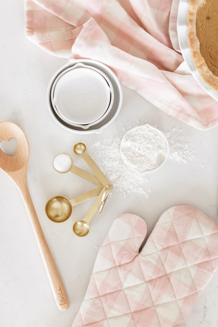 Utensílios de confeitaria vistos de cima: colher de madeira, medidores dourados, farinha e pano xadrez rosé